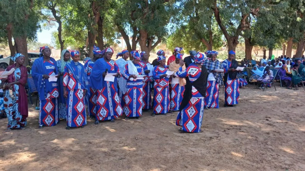 Un grupo de personas vestidas con coloridos trajes tradicionales con patrones de diamantes azules, rojos y blancos están en fila al aire libre, con árboles que proporcionan sombra en el fondo.