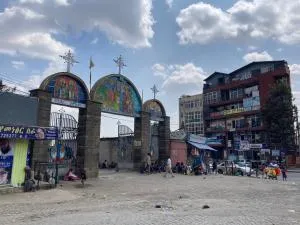 Una escena callejera con una entrada arqueada que presenta tres arcos rematados con cruces, coloridas obras de arte religioso, personas sentadas y de pie alrededor, y edificios en el fondo bajo un cielo parcialmente nublado.