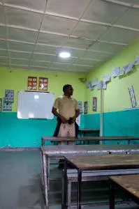 Una persona está de pie en un aula con paredes verdes, una pizarra y escritorios dispuestos en filas.