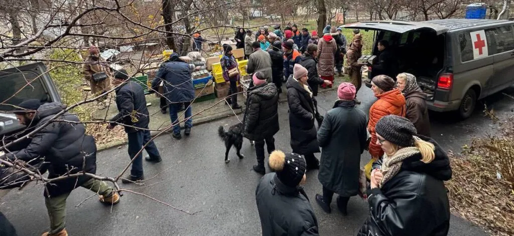 Un grupo de personas con ropa de invierno reunidas alrededor de una furgoneta con un símbolo de cruz roja, distribuyendo suministros al aire libre.